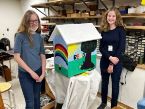 the girls who painted the little free library pose next to it in the workshop