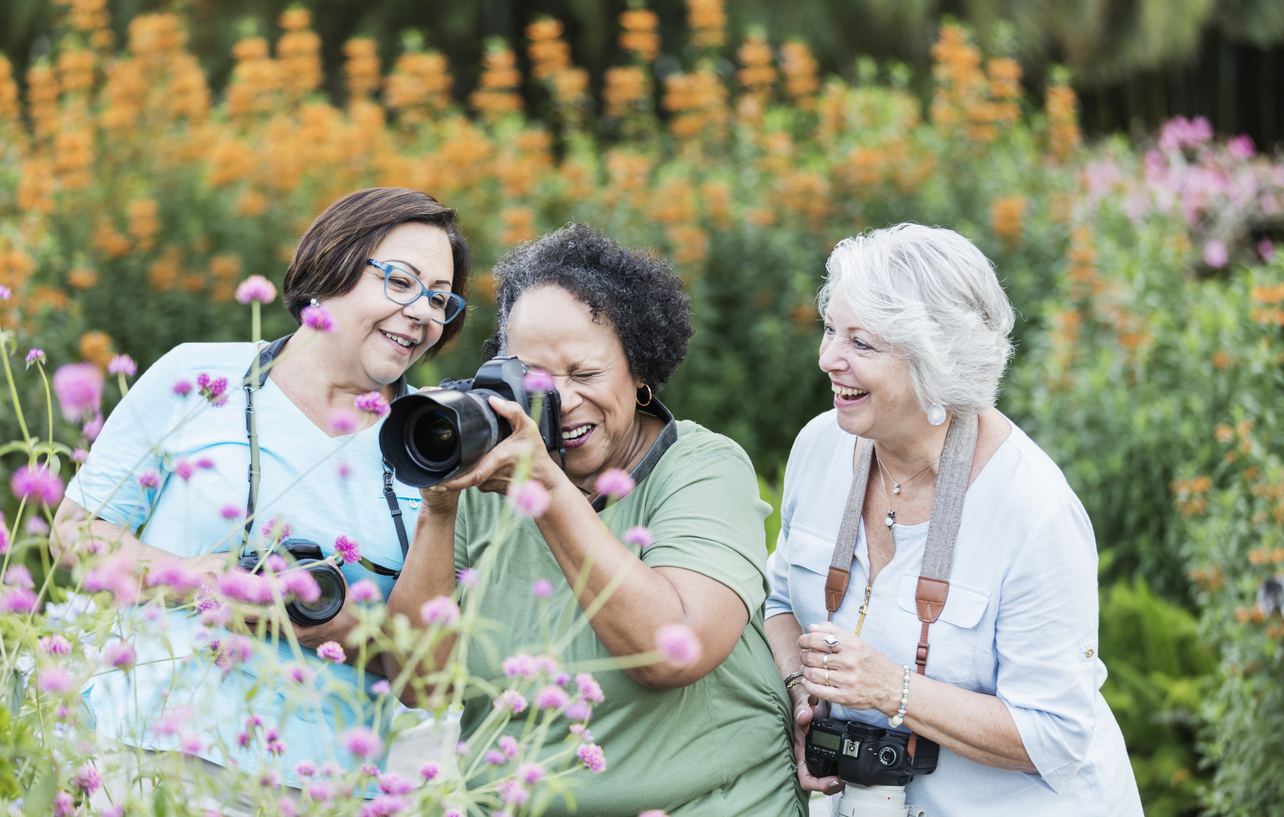 three older women with cameras outside. woman in the center is photographing a plant while her friends look on.