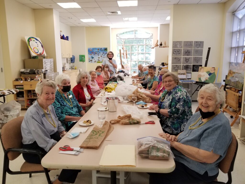 A large group of older adults are seated around an arts table. On the table are plastic bags. The bags are used to make mats for people without homes