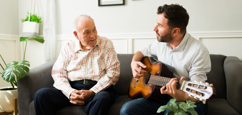 hospice volunteers. a young man plays guitar for an older man as they sit on a couch.