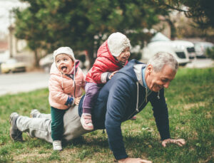 smiling older man with children playing on his back as he does a push up outdoors
