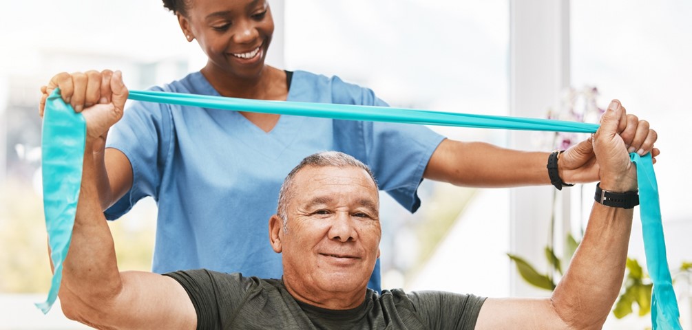 outpatient therapy. a physical therapist helps an older adult man with an exercise using a teal colored stretch band.