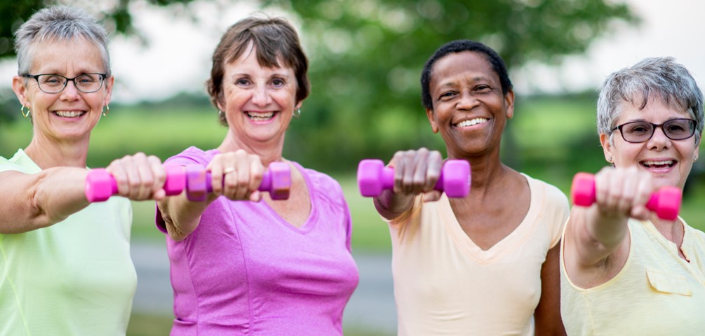 strength training. four older women pose outside holding pink hand weights