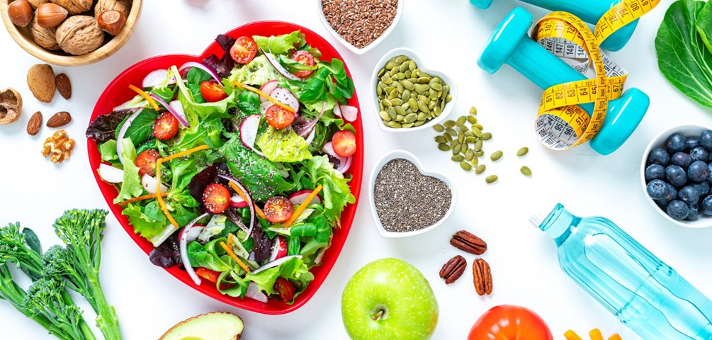 American Heart Month Small Steps to Protect Our Hearts. A vibrant salad in a red, heart-shaped bowl surrounded by nuts, seeds, a green apple, blue water bottle, blue hand weights and a yellow tape measurer.