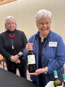 an older woman in black stands behind another older woman in blue who is holding up a wine bottle