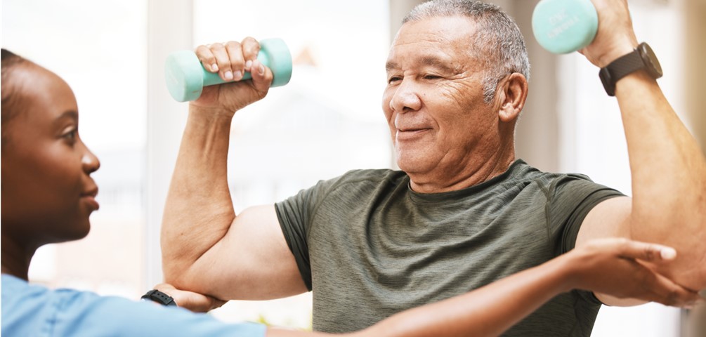 stroke rehabilitation. young woman in blue scrubs holds up arms of older man who is holding light blue dumbbells.