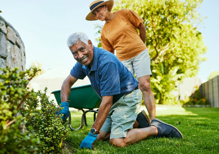 older man kneeling as he gardens, while older woman in yellow shirt and hat stands behind him