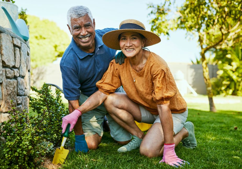 older man in blue shirt with older woman in yellow shirt and hat gardening together smiling at the camera