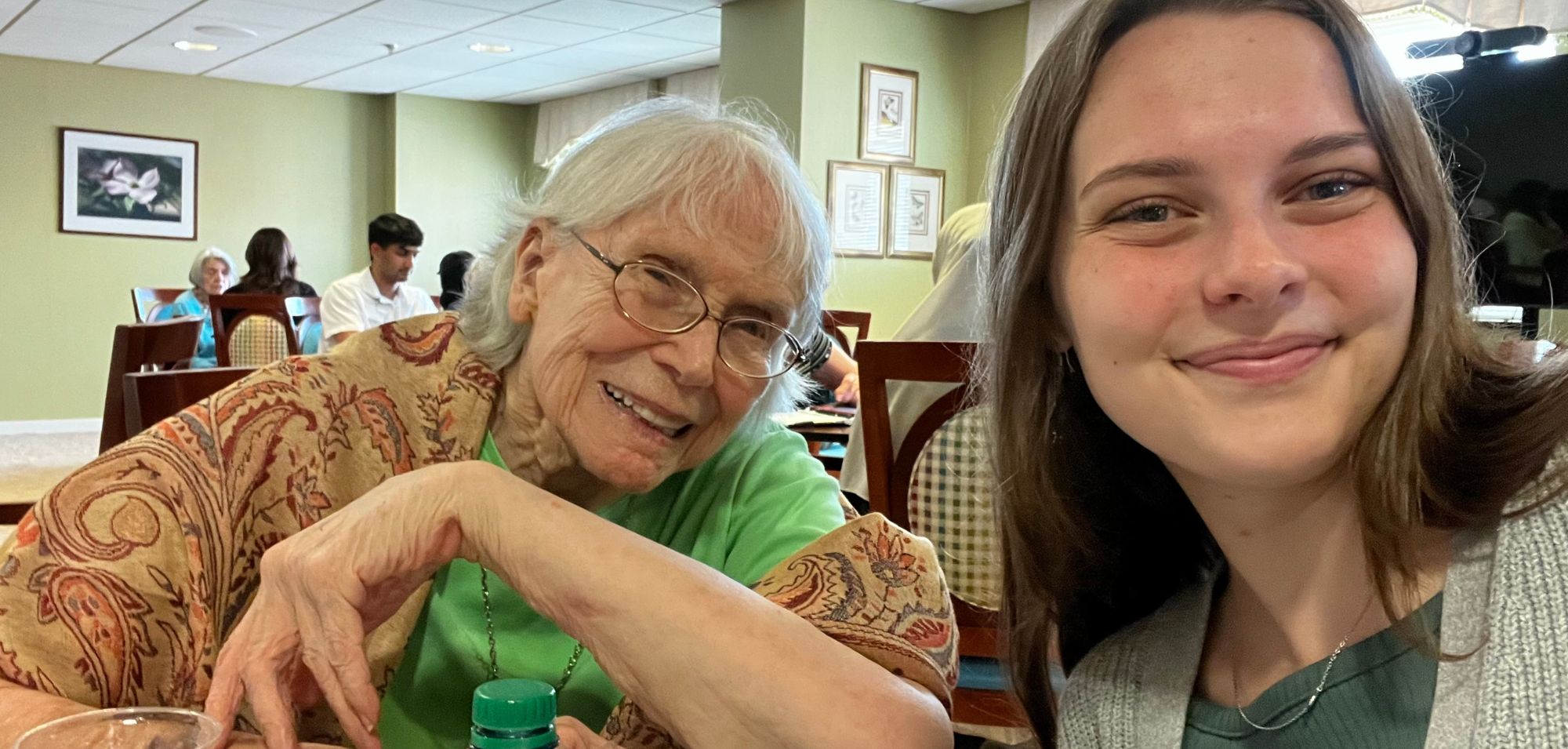 Resident with Parkinson’s Disease. Older woman in green shirt and orange shawl smiles next to young woman.