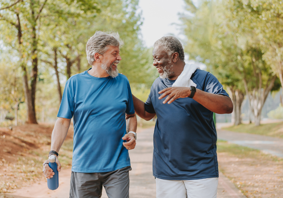 two older adult men talking as they exercise outside. The man on the left holds a water bottle, and the man on the right has a towel around his neck.