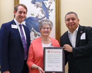 Inspired by her family history, Goodwin Living resident Rita Siebenaler sought support to launch the citizenship program. Here, she shares a joyful moment with Goodwin Living President and CEO Rob Liebreich and Mason District Supervisor Adres Jimenez as they proudly present a resolution of support from the Fairfax County Board of Directors during the ceremony.