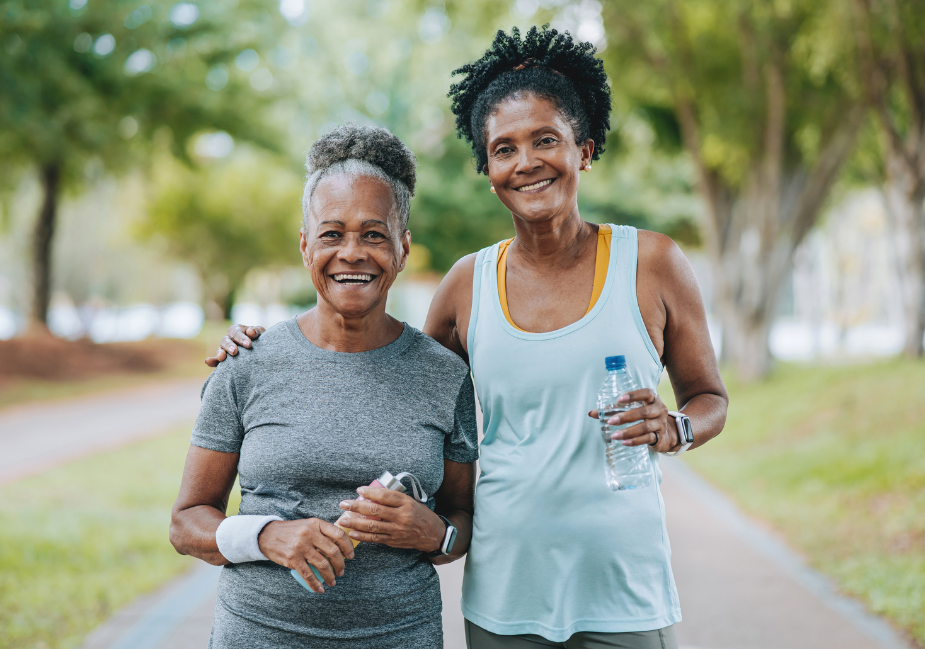 Two older adult women smile at the camera while exercising outside.