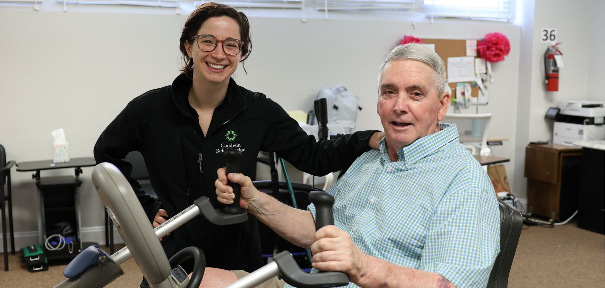 Vitala. Goodwin Rehabilitation therapist stands with a resident who is on an exercise bike. both are smiling at the camera.