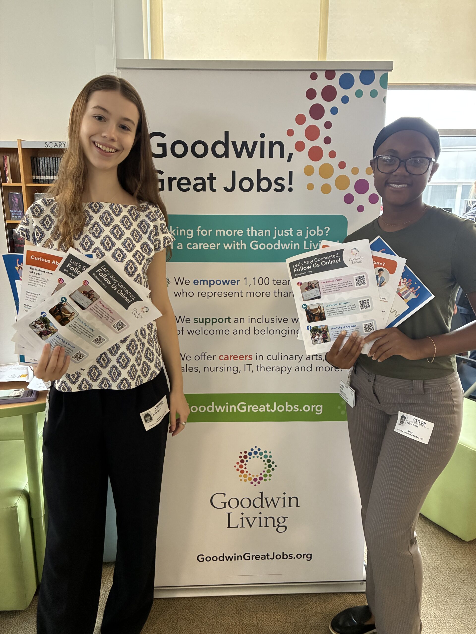 Photo: Marketing and Communications Intern Julia Carey and Human Resources Intern Atiya Idris hold handouts as they stand in front of a Goodwin Living banner at an Alexandria City Public Schools summer career fair.