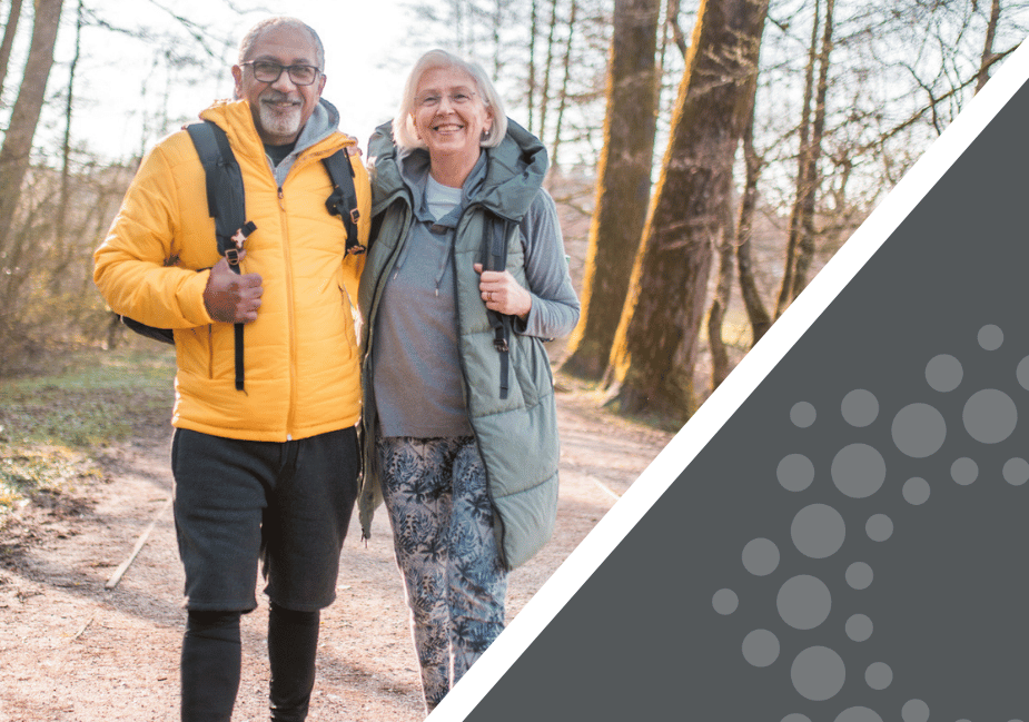 senior couple wearing winter coats and backpacks pose outside as they take a walk in the woods