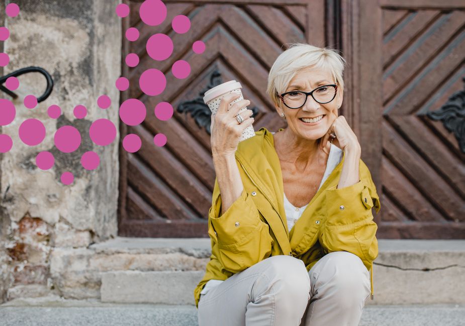 older woman in a yellow top sits on steps in front of ornate wooden doors, holding a coffee cup and smiling at the camera