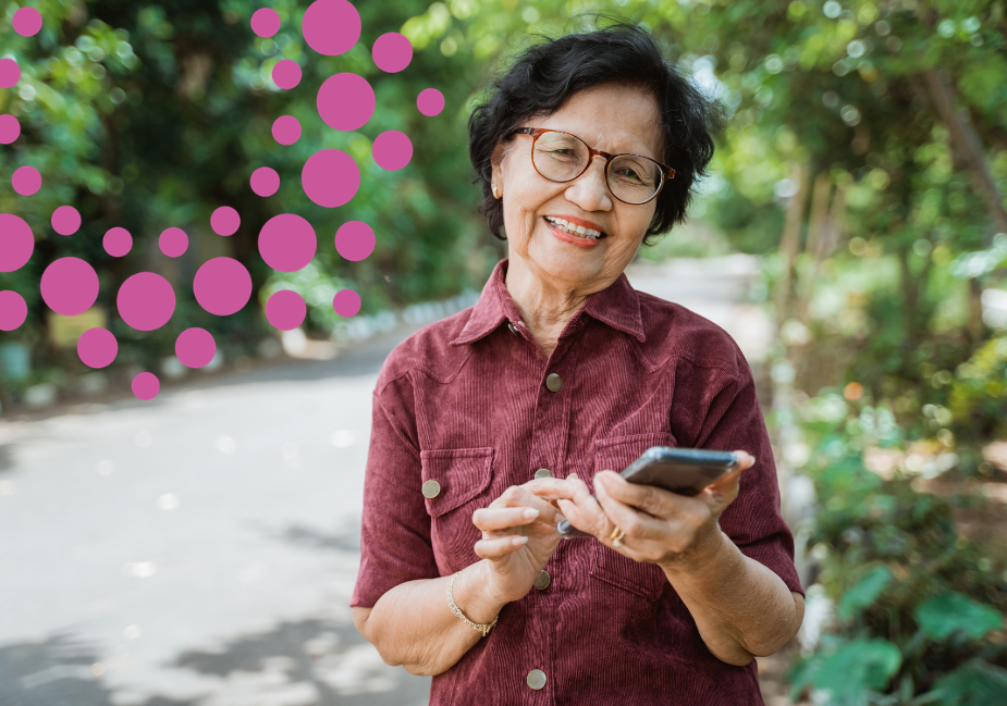 an older Asian woman smiles at the camera as she stands outside holding a smartphone