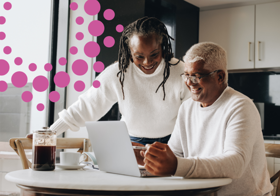 older man in white sweater sits smiling at a computer. older woman in white sweater stands over his shoulder smiling down at the computer